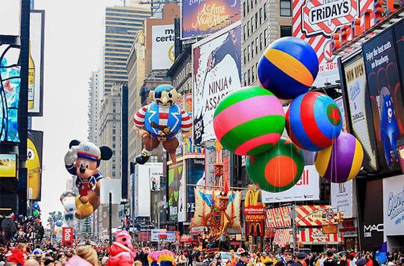 Grandi e colorati palloni da parata, incluso Topolino, sfilano lungo una strada cittadina affollata tra alti edifici con molti cartelloni pubblicitari.