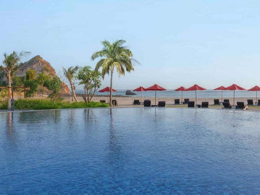 Ein großer Infinity-Pool überblickt einen Sandstrand mit roten Sonnenschirmen, Liegestühlen und Palmen unter klarem Himmel.