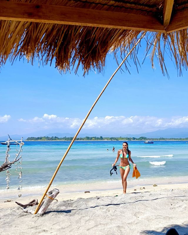 Eine Frau im Bikini mit Schnorchelausrüstung geht an einem Sandstrand mit türkisfarbenem Wasser, von einem Strohdach aus gesehen.