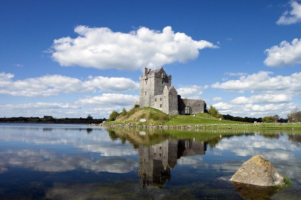 Un castello di pietra su una riva erbosa si riflette nell'acqua calma del lago sotto un cielo azzurro parzialmente nuvoloso.