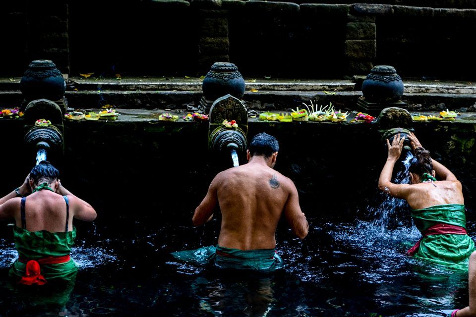 Un voyage de groupe WeRoad vu de dos participant à un rituel de purification sous des fontaines d'eau en pierre dans un bassin sombre.