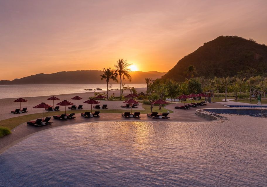 Ein Resort-Pool blickt auf einen Sandstrand mit Liegestühlen und roten Sonnenschirmen, während die Sonne über dem Meer und den Hügeln untergeht.