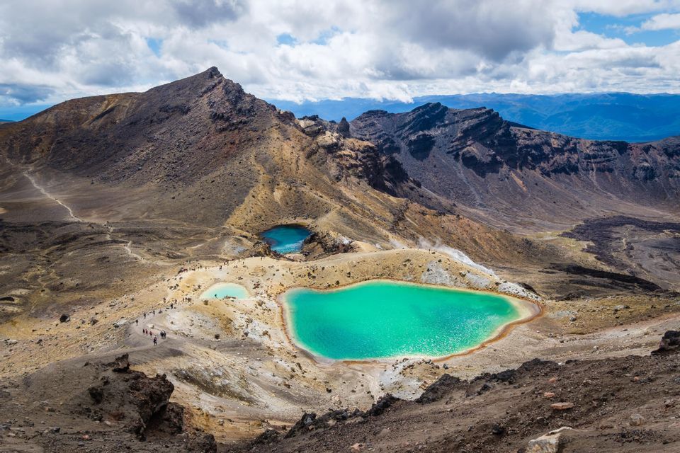A WeRoad group trip hikes on a trail overlooking turquoise volcanic crater lakes in a mountainous landscape under a cloudy sky.