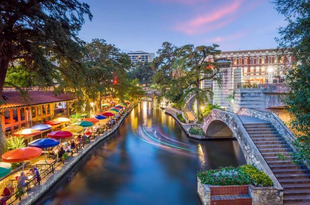 Un canale fluviale al tramonto con persone che cenano sotto ombrelloni colorati sulla riva, un ponte di pietra e scie luminose da una barca.