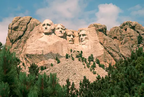 Il monumento del Monte Rushmore scolpito in una montagna di granito, con pini verdi in primo piano e un cielo azzurro sopra.