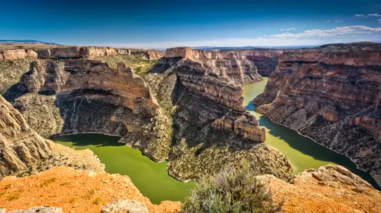 Un fiume verde si snoda in un vasto canyon roccioso sotto un cielo azzurro, visto da un punto panoramico elevato.