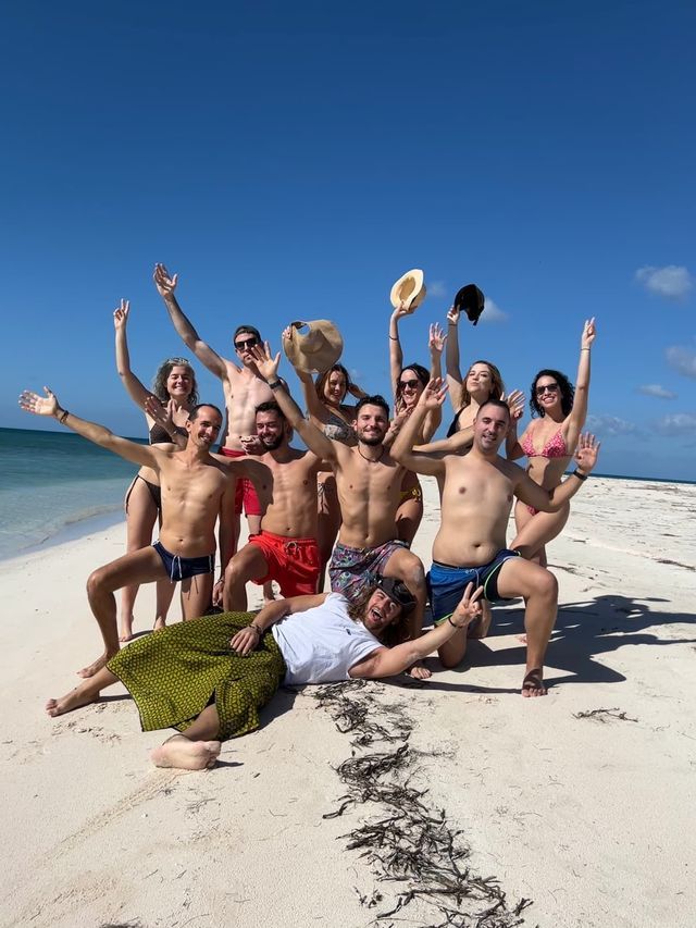 Eine WeRoad-Gruppe lächelt und posiert für ein Foto an einem weißen Sandstrand unter klarem Himmel.