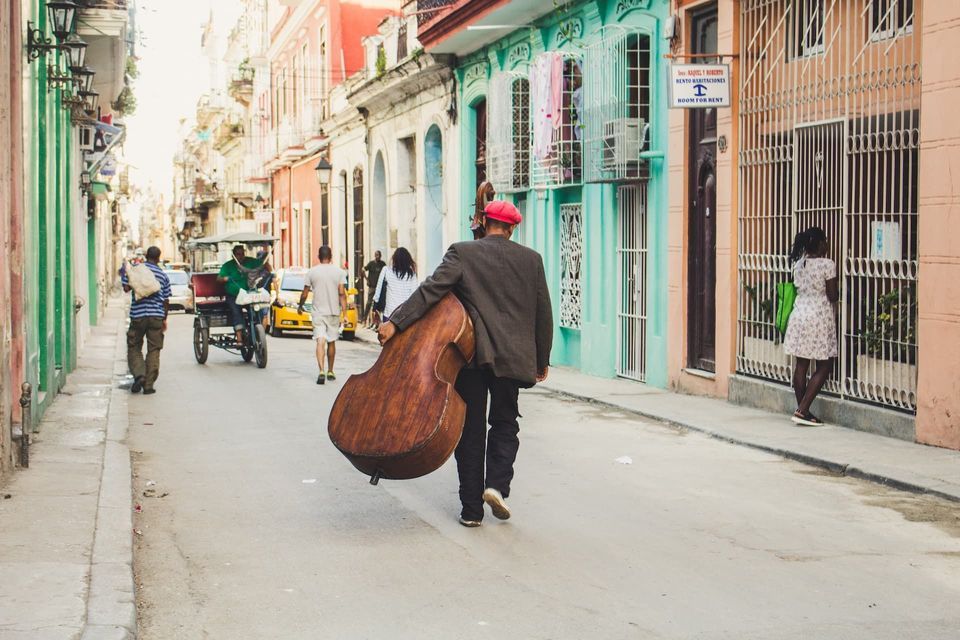Un hombre con gorra roja, visto de espaldas, lleva un contrabajo mientras camina por una calle estrecha bordeada de edificios coloridos.