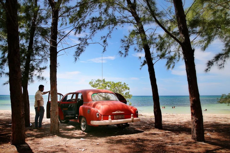 Une voiture rouge vintage, portes ouvertes, est garée sous les pins sur une plage de sable, un homme se tient à côté.