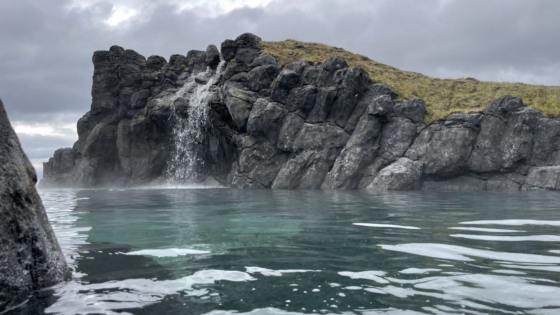 Ein kleiner Wasserfall stürzt über eine dunkle Felsformation in das dampfende Wasser einer natürlichen heißen Quelle.