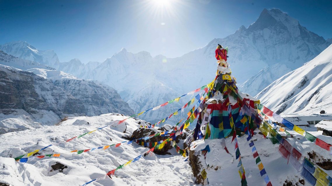 Coloridas banderas de oración adornan un santuario de roca cubierto de nieve frente a una vasta cordillera nevada bajo un sol brillante.