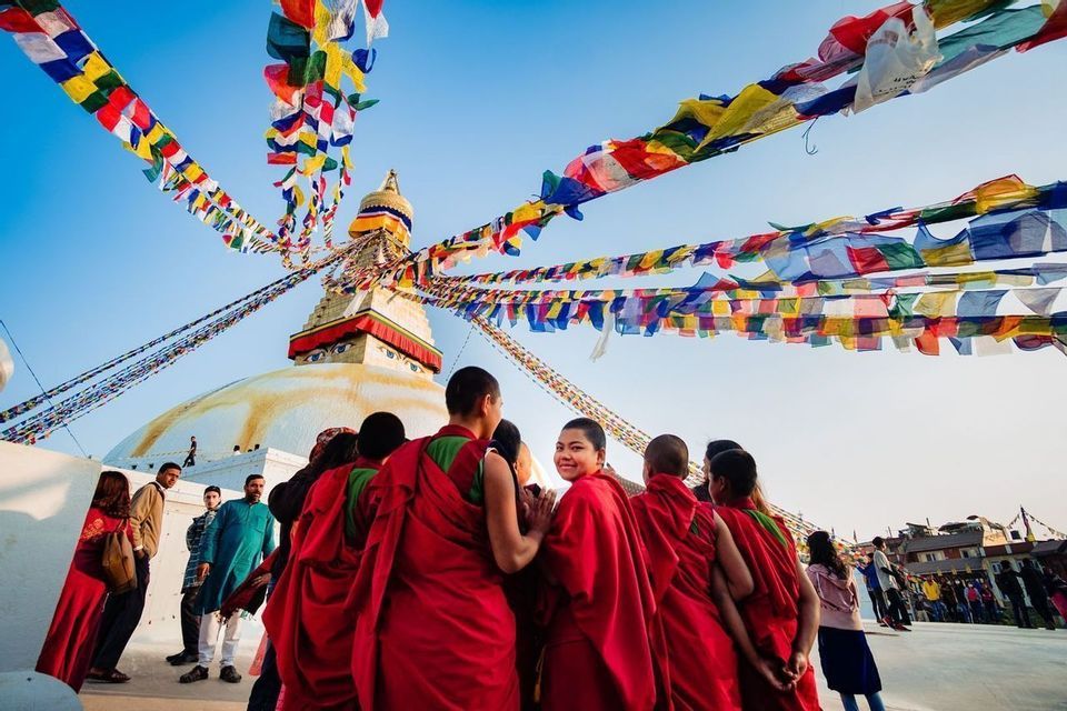 A group of people in red robes look towards a large white stupa with colorful prayer flags strung from its spire against a blue sky.