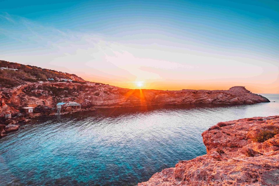 The setting sun casts an orange glow over red rocky cliffs surrounding a calm cove with clear blue water.
