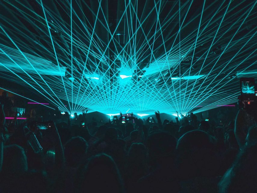 Una folla di sagome di persone in un locale buio sotto uno spettacolo di luci laser ciano vibranti e incrociate.