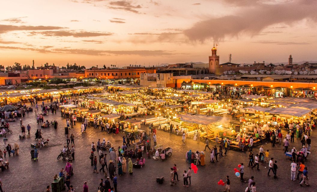 An elevated view of a bustling market square at sunset, filled with people walking between brightly lit stalls.