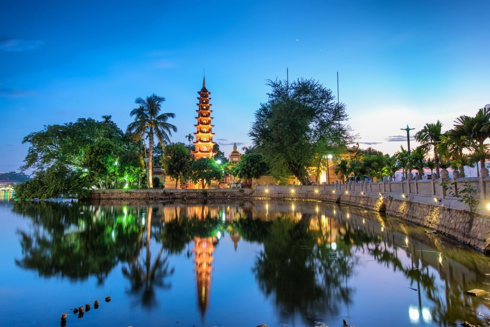 Una torre pagoda iluminada y árboles circundantes reflejándose en el agua en calma al anochecer.