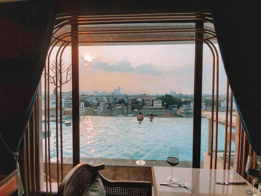 Deux personnes dans une piscine à débordement sur un toit, surplombant un paysage urbain au coucher du soleil, vues depuis une chambre avec un verre de vin sur une table.