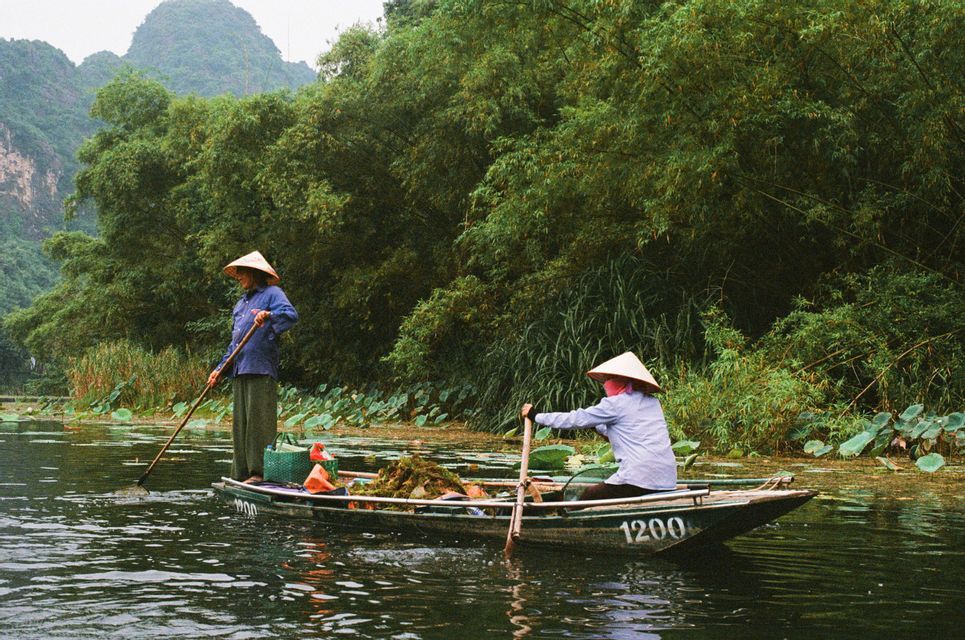 Due persone con cappelli conici navigano su una barca lungo un fiume, immerse tra vegetazione rigogliosa e colline, una manovrando con una pertica e l'altra remando.