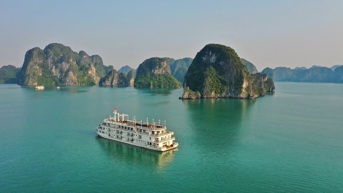 A large white cruise ship sails on calm turquoise water among towering limestone karsts covered in greenery.