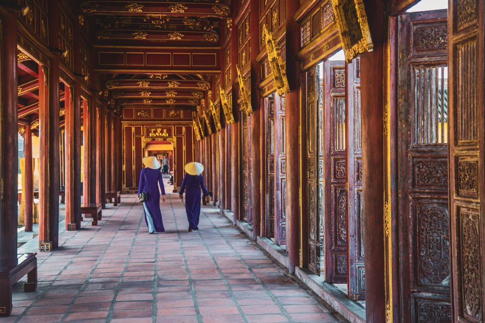 Due donne con cappelli conici e abiti tradizionali viola camminano lungo un lungo e ornato corridoio con pilastri di legno rossi e dorati.