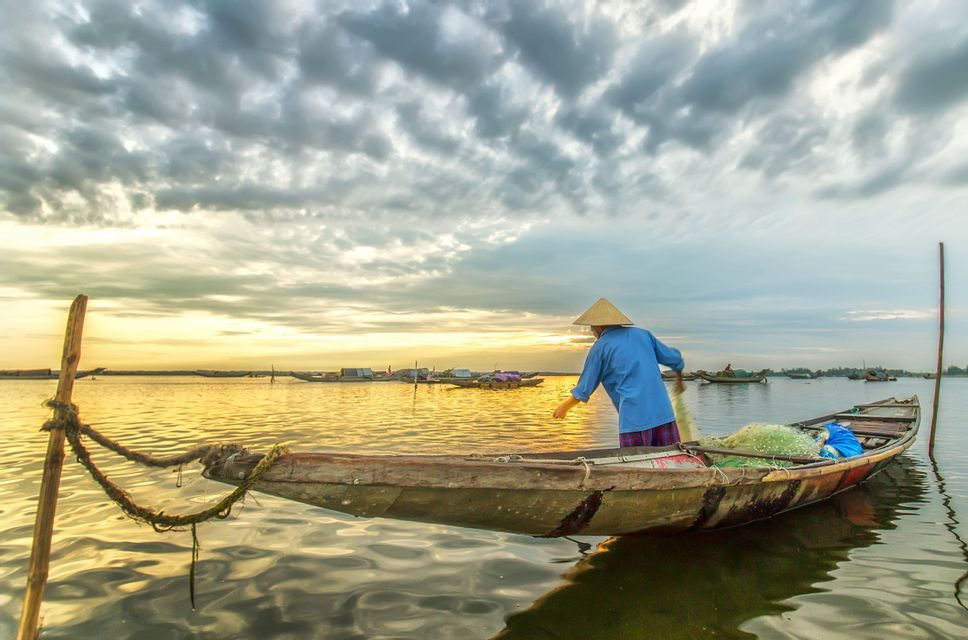 Un pêcheur coiffé d'un chapeau conique manipule un filet de pêche sur une petite barque en bois au coucher du soleil sous un ciel nuageux.