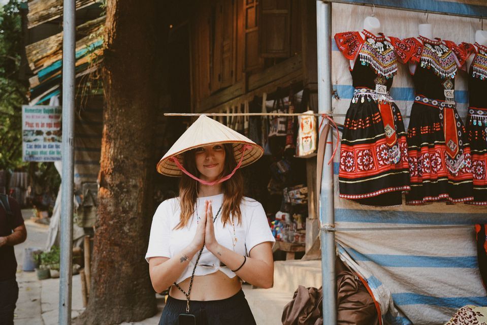 Una donna con un cappello conico sorride con le mani giunte davanti a una bancarella del mercato che espone abiti tradizionali.