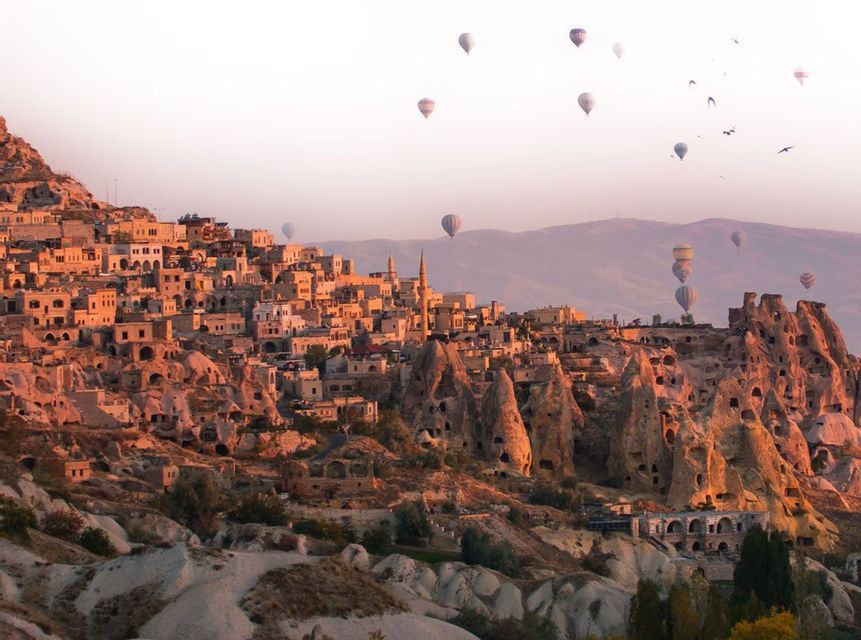 Des montgolfières flottent au-dessus d'un village construit dans des falaises rocheuses et des habitations troglodytes lors d'un lever de soleil brumeux.