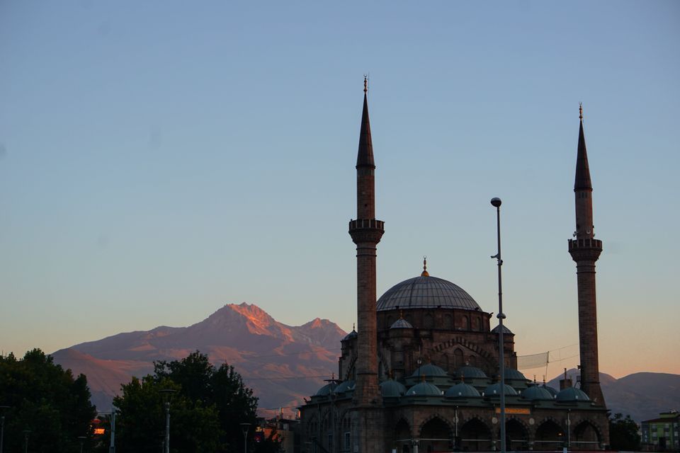 Une grande mosquée avec deux minarets et plusieurs dômes se dresse devant une chaîne de montagnes illuminée par le soleil sous un ciel clair.