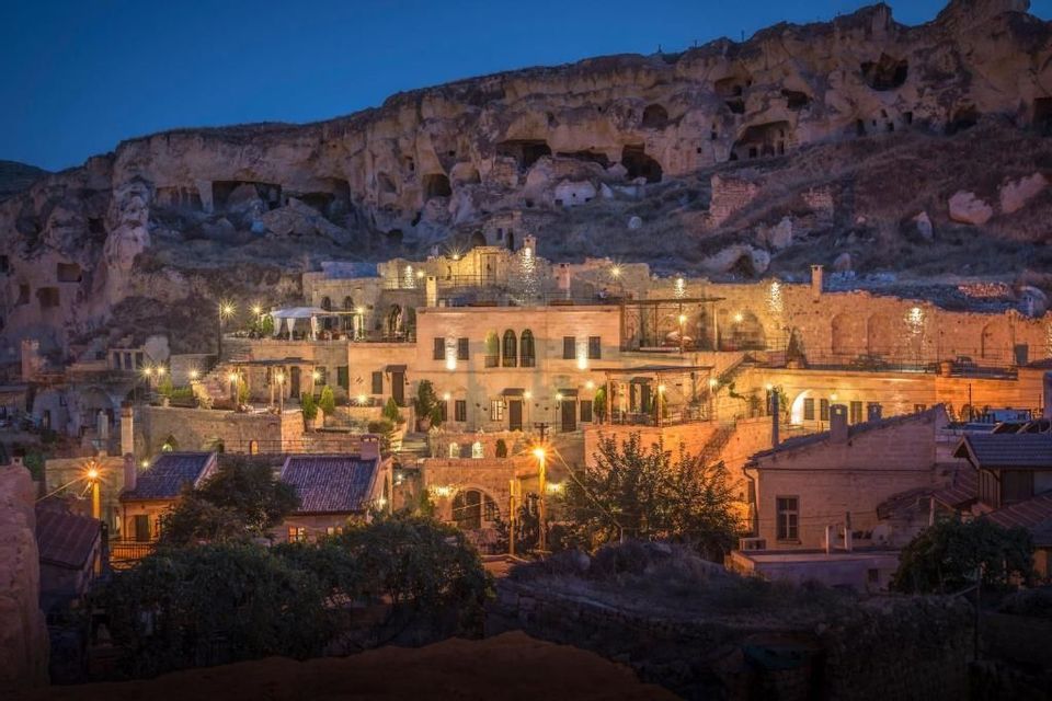 Edifici in pietra illuminati e abitazioni rupestri incastonate in un fianco roccioso al crepuscolo.
