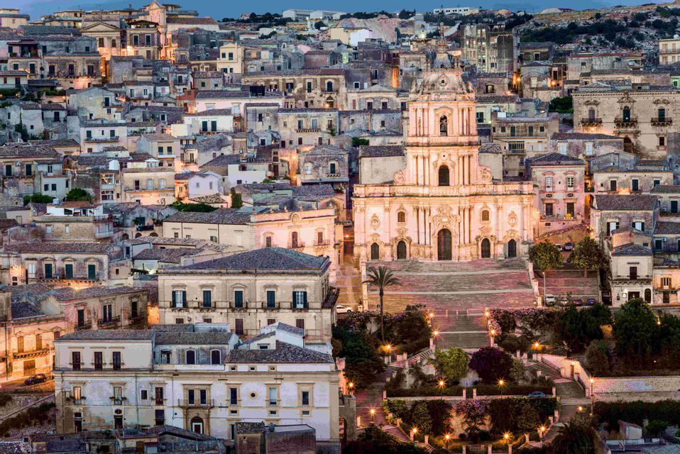 An illuminated baroque church with a grand staircase sits in the middle of a dense hillside city at dusk.