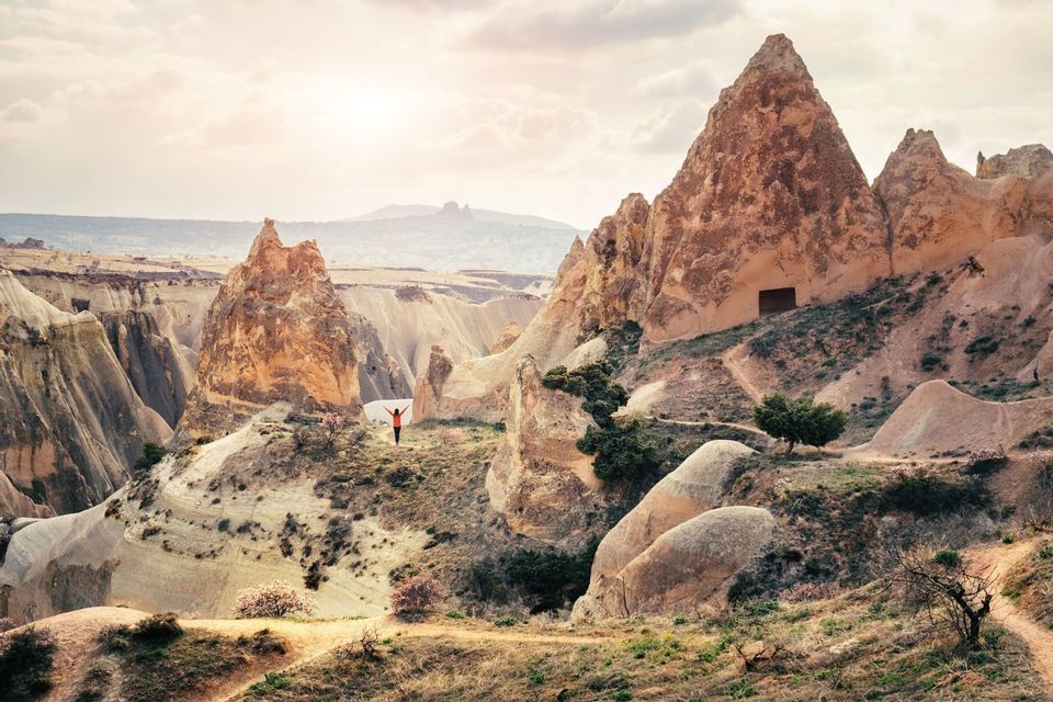 Una persona sola con un top arancione sta in piedi con le braccia alzate, affacciata su una valle rocciosa con formazioni coniche e abitazioni rupestri.