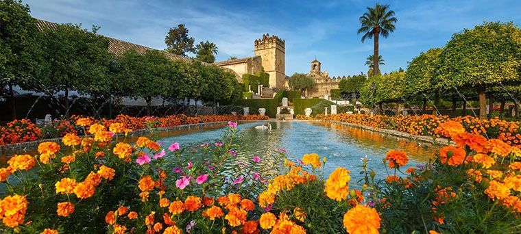 A long reflecting pool in a formal garden is flanked by orange marigolds and trees, with a historic stone building in the background.