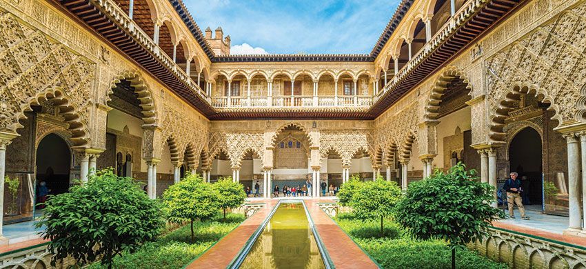 Un elaborato cortile moresco con una vasca centrale riflettente, giardini e archi finemente intagliati sotto un cielo blu.