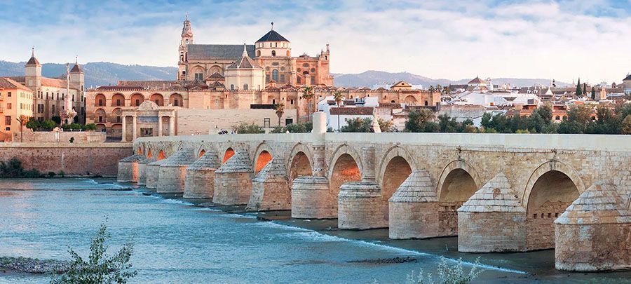 A historic stone bridge with many arches crosses a river towards a city with ancient buildings at sunset.