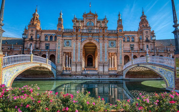 An ornate brick building with tiled bridges is reflected in a canal, with pink flowers in the foreground.