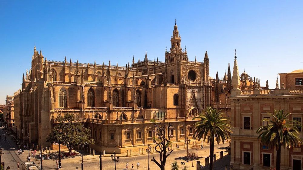 Una cattedrale gotica riccamente decorata con un'alta torre campanaria si staglia contro un cielo azzurro limpido sopra una via cittadina con palme.