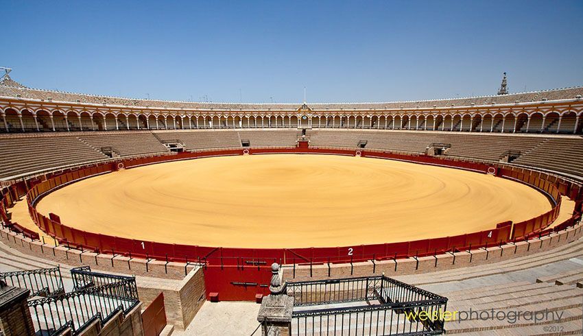 Ampia veduta di un'arena da corrida vuota con anello di sabbia dorata, gradinate e un colonnato ad arco sotto un cielo azzurro e limpido.
