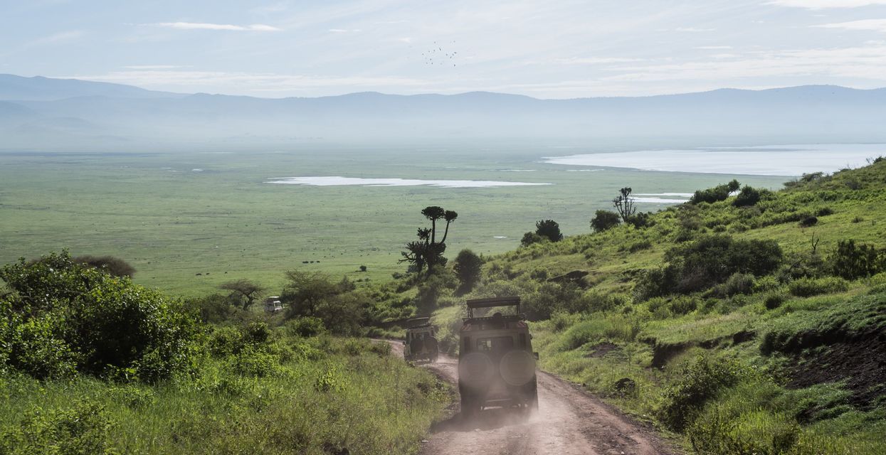 Deux jeeps de safari descendent un chemin de terre sinueux sur une colline herbeuse, surplombant une vaste vallée verdoyante avec un lac et des montagnes lointaines.