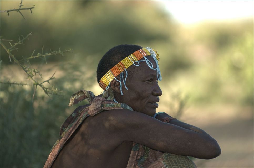 Eine ältere Frau im Profil, die ein traditionelles Perlenstirnband und Tuch trägt und mit verschränkten Armen im Freien sitzt.