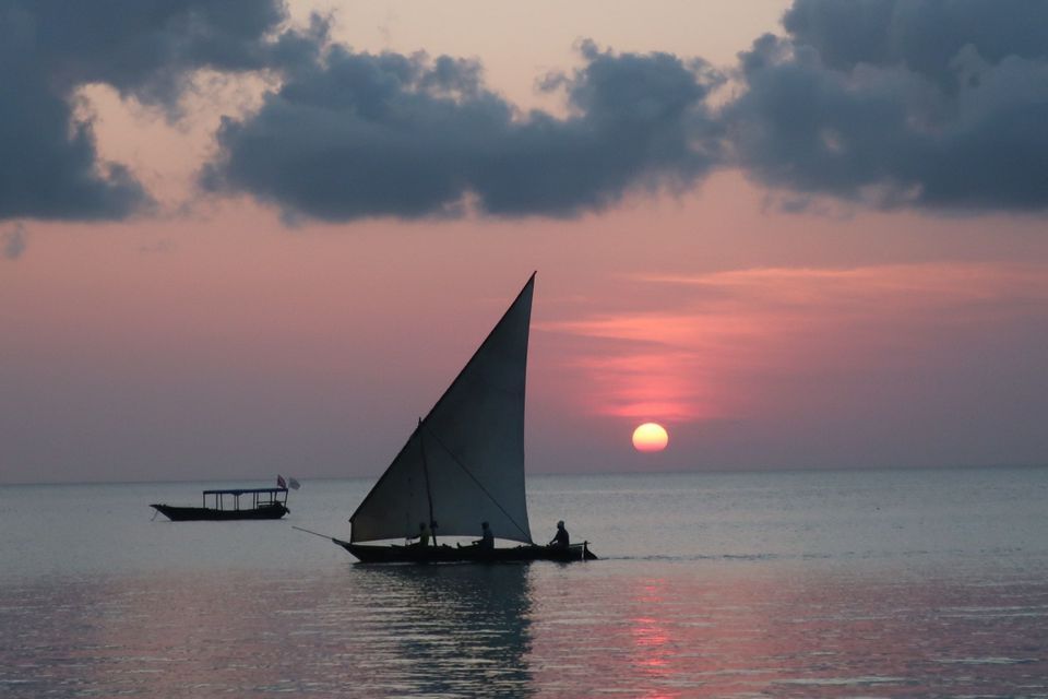 Un velero con personas a bordo se desliza sobre aguas tranquilas al atardecer, con el sol bajo en el horizonte bajo un cielo rosado y nublado.