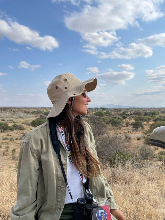 A woman on a WeRoad group trip wearing a safari hat and binoculars looks out over a savanna landscape.