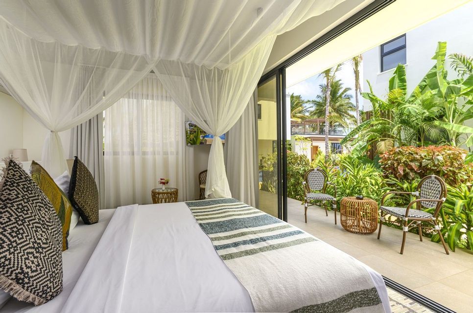 A bright bedroom with a white canopy bed overlooks a private patio with chairs and lush tropical plants.