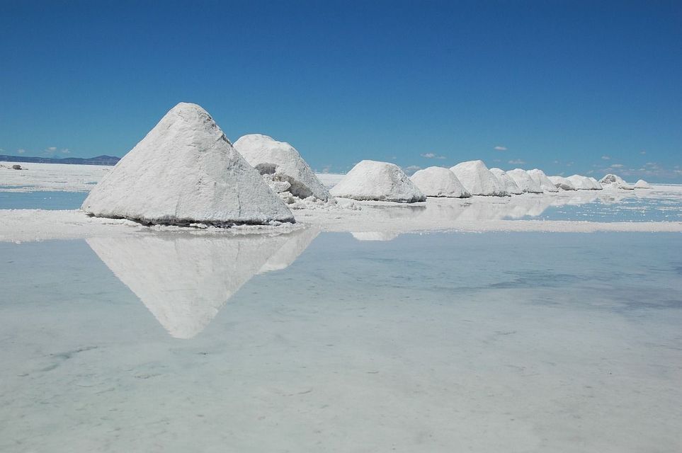 Una fila di cumuli conici di sale su una salina allagata si riflette nell'acqua calma sotto un cielo azzurro e sereno.