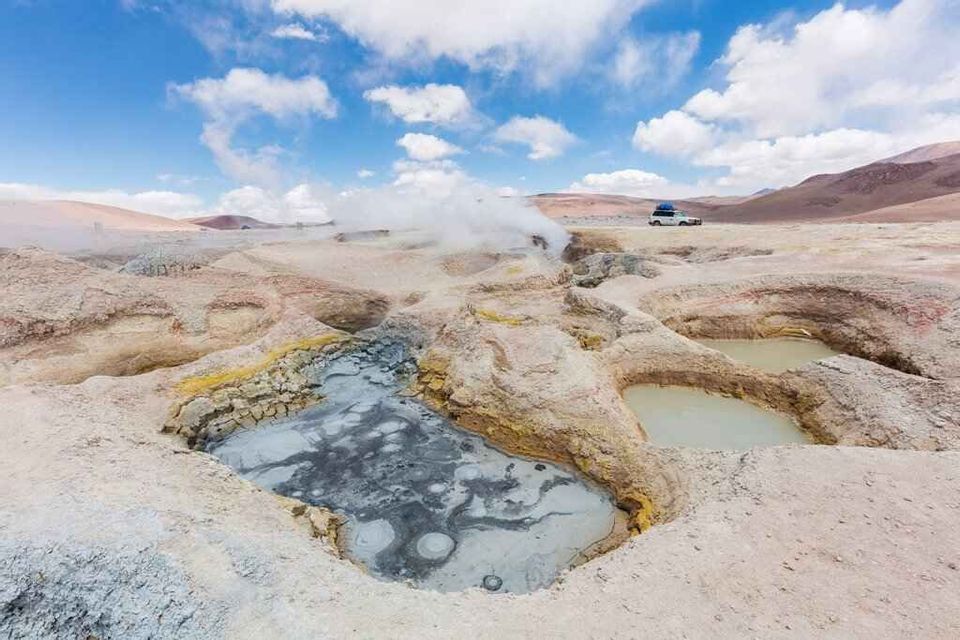 Pozze di fango ribollenti e fumarole vaporeggianti in un vasto campo geotermico, sotto un cielo blu e nuvoloso, con un veicolo in lontananza.