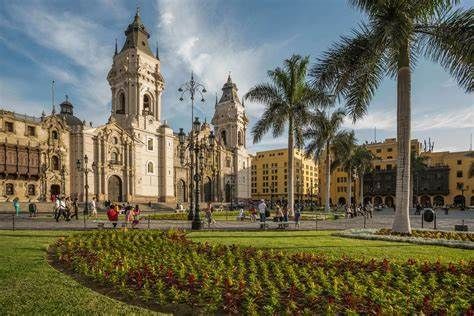 Una cattedrale storica con due campanili si affaccia su una piazza pubblica soleggiata con palme, aiuole fiorite e persone che passeggiano.