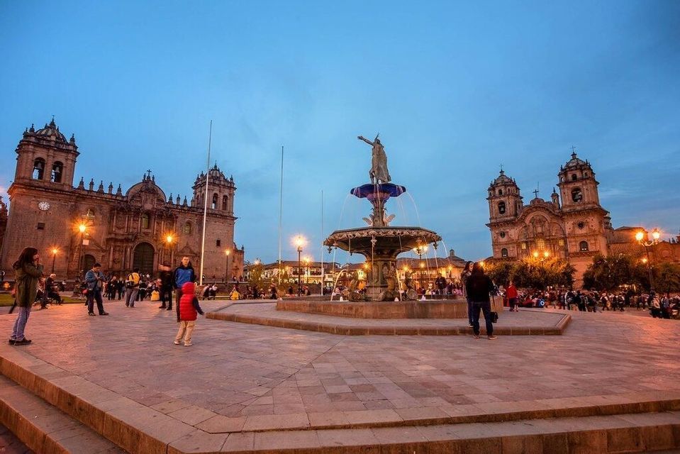 Una piazza storica al crepuscolo con un'elegante fontana centrale, affiancata da due cattedrali illuminate mentre le persone passeggiano.