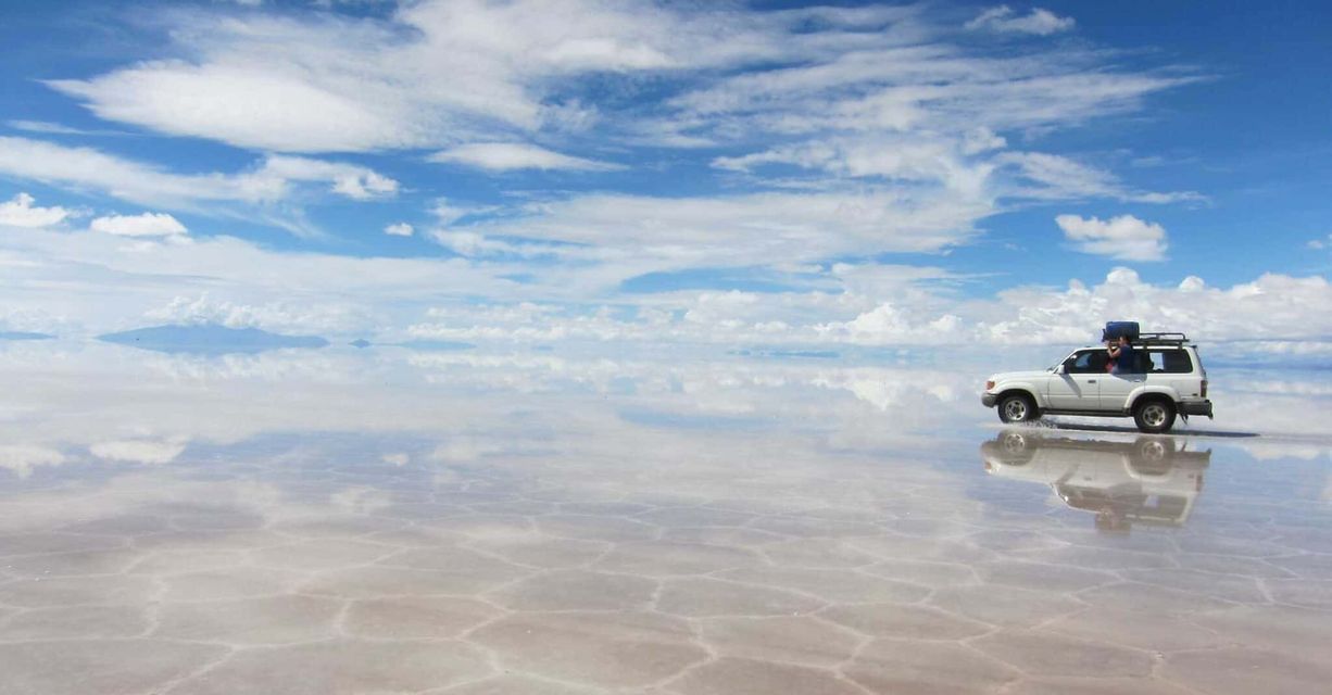 Un SUV bianco di un viaggio di gruppo WeRoad che attraversa una vasta e umida distesa di sale che riflette il cielo blu nuvoloso come uno specchio.