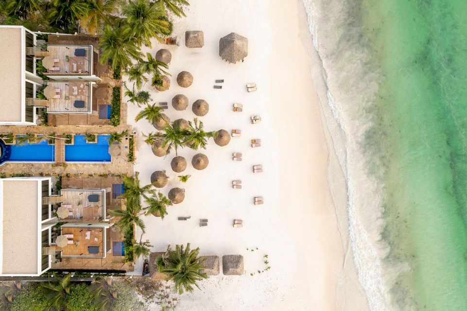 Luftaufnahme eines Resorts mit Pools und Palmen an einem weißen Sandstrand und türkisfarbenem Meer.
