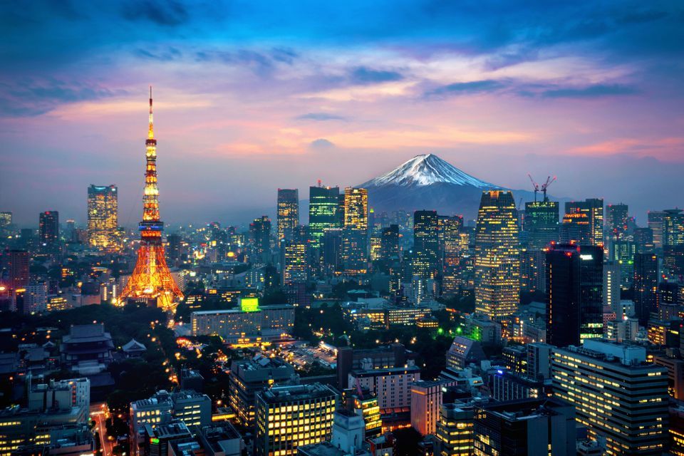 A city skyline at twilight, featuring an illuminated orange tower and a distant, snow-capped mountain under a colorful sky.