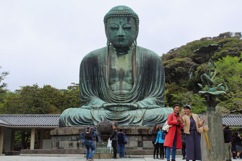 Une immense statue de Bouddha en bronze, assise, surplombe les touristes prenant des photos dans une cour extérieure.